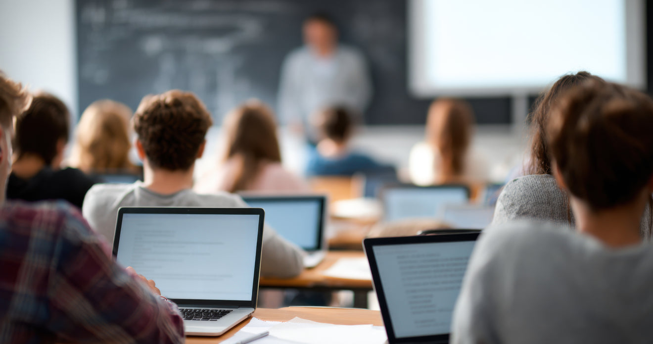 Students using laptops in a classroom while a teacher instructs from the front of the room.
